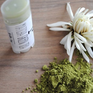 K&ouml;nnte beinhalten: Eine wei&szlig;e Plastikr&ouml;hre mit einem Etikett mit der Aufschrift "Matcha Green Tea Deodorant" und einem gr&uuml;nen Deodorantstift im Inneren. Ein Haufen gr&uuml;nes Matcha-Pulver und eine wei&szlig;e Blume liegen auf einer Holzoberfl&auml;che.