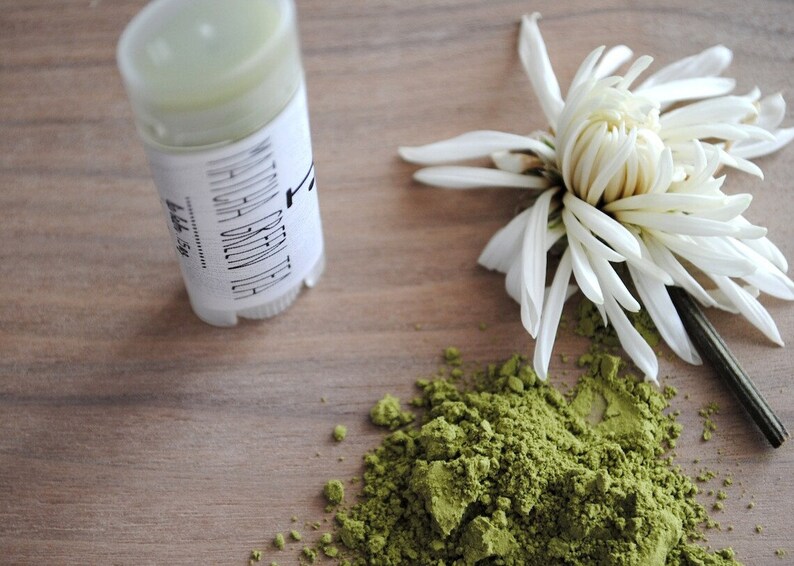 K&ouml;nnte beinhalten: Eine wei&szlig;e Plastikr&ouml;hre mit einem Etikett mit der Aufschrift "Matcha Green Tea Deodorant" und einem gr&uuml;nen Deodorantstift im Inneren. Ein Haufen gr&uuml;nes Matcha-Pulver und eine wei&szlig;e Blume liegen auf einer Holzoberfl&auml;che.