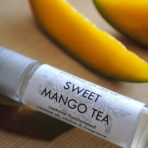 May include: A small, clear glass bottle with a white label that reads "perfume oil .35 oz SWEET MANGO TEA contains: fractionated coconut oil, vitamin E, fragrance". The bottle is sitting on a wooden surface next to two slices of mango.