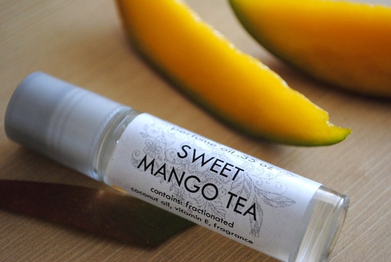 May include: A small, clear glass bottle with a white label that reads "perfume oil .35 oz SWEET MANGO TEA contains: fractionated coconut oil, vitamin E, fragrance". The bottle is sitting on a wooden surface next to two slices of mango.