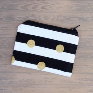 May include: A small, rectangular zippered pouch with a black and white striped pattern. The pouch features three gold-colored polka dots and a black zipper. The pouch is likely used for carrying small items like cosmetics or accessories.