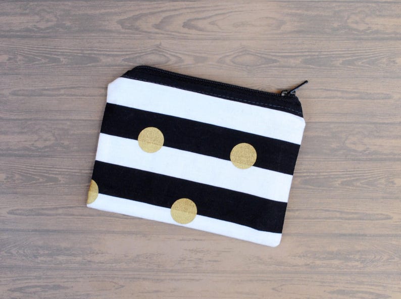 May include: A small, rectangular zippered pouch with a black and white striped pattern. The pouch features three gold-colored polka dots and a black zipper. The pouch is likely used for carrying small items like cosmetics or accessories.