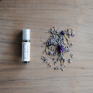 May include: A small glass bottle with a silver roller ball top. The bottle is labelled "French Lavender" and contains a clear liquid. Dried lavender flowers are scattered around the bottle on a wooden surface.