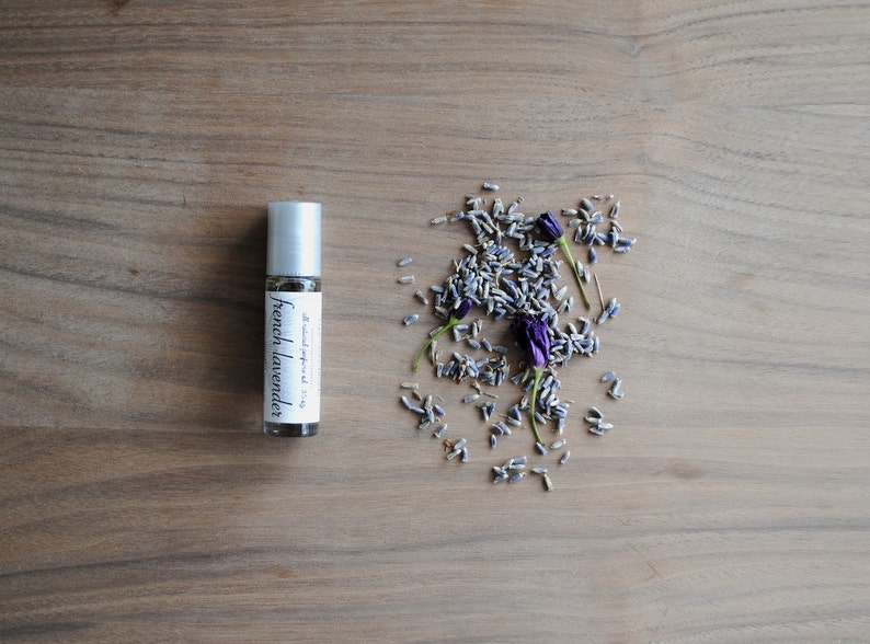 May include: A small glass bottle with a silver roller ball top. The bottle is labelled "French Lavender" and contains a clear liquid. Dried lavender flowers are scattered around the bottle on a wooden surface.