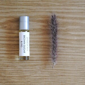 May include: A perfume oil bottle with a silver cap and a label that reads "BROWN SUGAR & FIG". The bottle is next to a dried, feathery plant stem. The background is a light brown wooden surface. The bottle contains .35 oz of perfume oil.