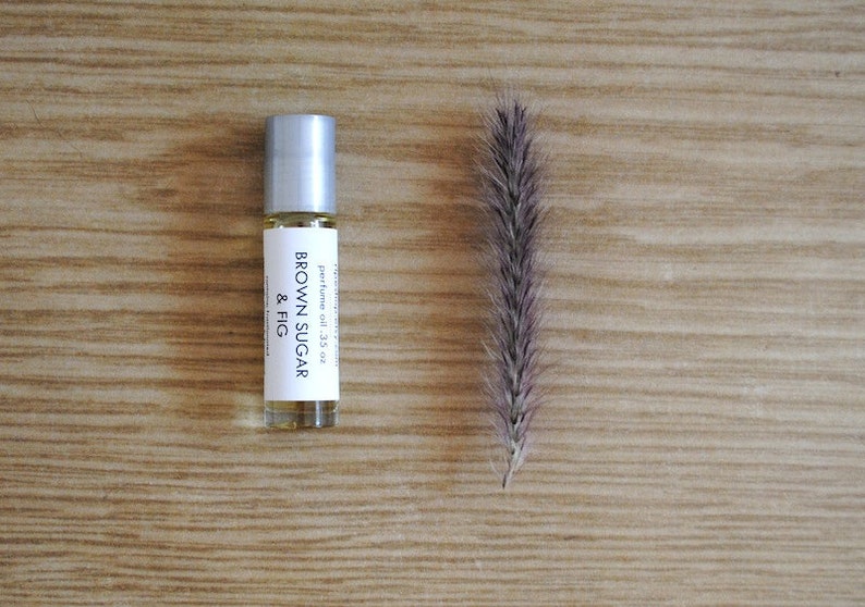May include: A perfume oil bottle with a silver cap and a label that reads "BROWN SUGAR & FIG". The bottle is next to a dried, feathery plant stem. The background is a light brown wooden surface. The bottle contains .35 oz of perfume oil.