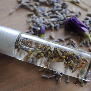 May include: A clear glass roller bottle filled with dried lavender buds. The bottle has a silver cap and is lying on a wooden surface.