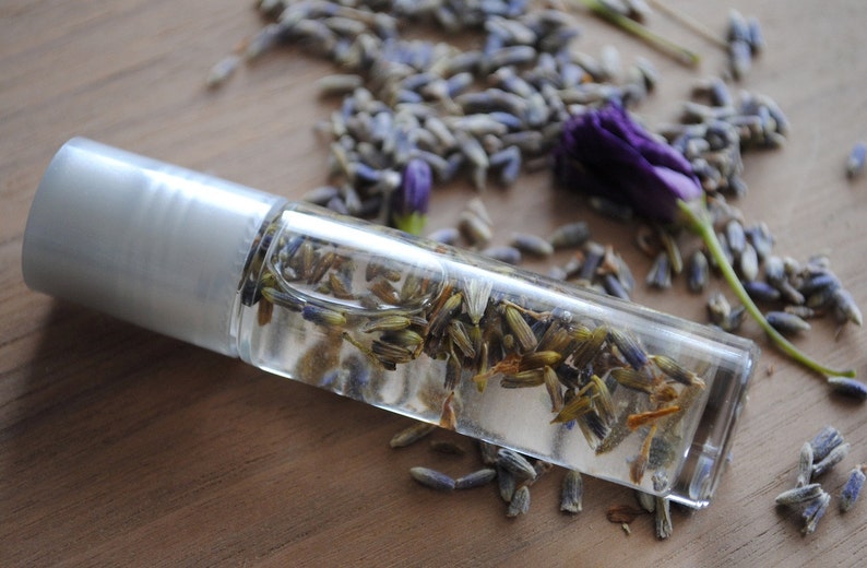 May include: A clear glass roller bottle filled with dried lavender buds. The bottle has a silver cap and is lying on a wooden surface.