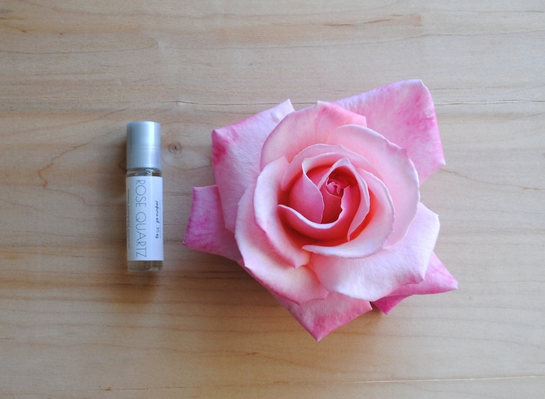 perfume oil in glass rollerball bottle in rose quartz sitting next to a large pink rose bloom