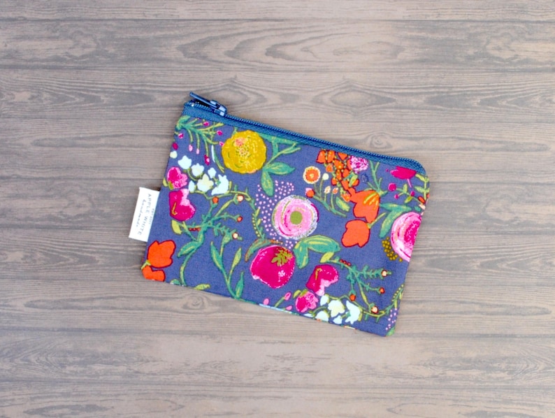 May include: A floral print pouch with a blue zipper. The pouch features a pattern of pink, orange, and yellow flowers on a blue background. A small white tag is attached to the side. The pouch is rectangular in shape.