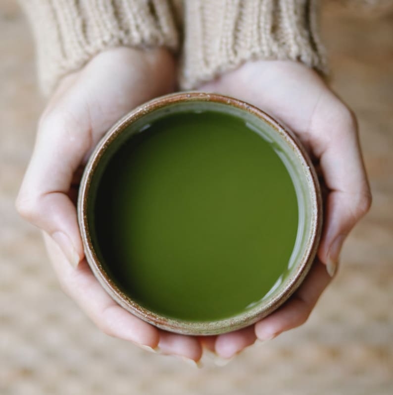 K&ouml;nnte beinhalten: Zwei H&auml;nde halten eine braune Keramikschale gef&uuml;llt mit leuchtend gr&uuml;nem Matcha-Tee.