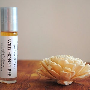 May include: A clear glass roll-on bottle of "Wild Honey Bee" perfume oil, next to a decorative cream-colored flower. The bottle has a silver cap and a white label with product information. The perfume oil is .35 oz.