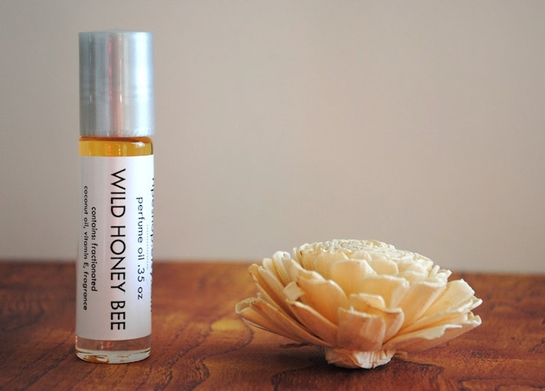 May include: A clear glass roll-on bottle of "Wild Honey Bee" perfume oil, next to a decorative cream-colored flower. The bottle has a silver cap and a white label with product information. The perfume oil is .35 oz.