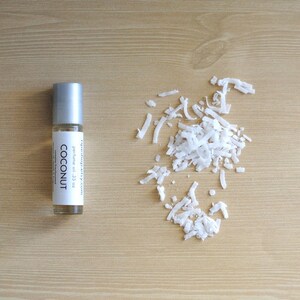 Puede incluir: Un peque&ntilde;o frasco de aceite de perfume de coco con rodillo plateado. El frasco est&aacute; etiquetado con "COCONUT" y "perfume oil .33 oz". El frasco est&aacute; sobre una superficie de madera marr&oacute;n claro con un mont&oacute;n de copos de coco blancos.