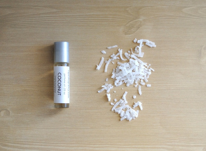 Puede incluir: Un peque&ntilde;o frasco de aceite de perfume de coco con rodillo plateado. El frasco est&aacute; etiquetado con "COCONUT" y "perfume oil .33 oz". El frasco est&aacute; sobre una superficie de madera marr&oacute;n claro con un mont&oacute;n de copos de coco blancos.