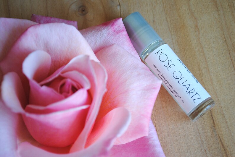May include: A small clear glass bottle with a white label that says "ROSE QUARTZ" and "contains fractionated coconut oil, vitamin E, fragrance". The bottle is sitting on a pink rose.