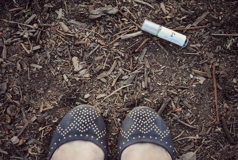 A glass rollerball perfume oil bottle laying on the ground with twigs and stones next to a pair of shoes.