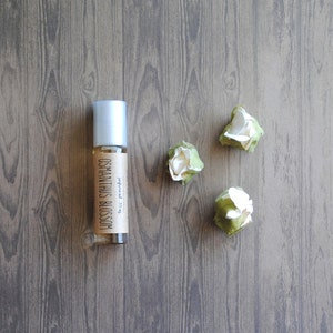 May include: A clear glass roller bottle with a silver cap and a label that reads "OSMANTHUS BLOSSOM." The bottle is next to three small, white and green artificial flowers. The items are on a wooden surface.