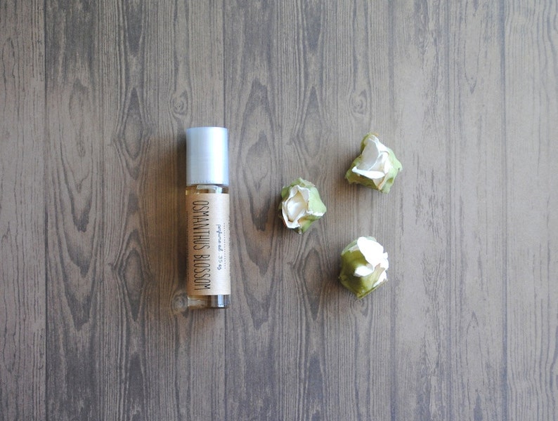 May include: A clear glass roller bottle with a silver cap and a label that reads "OSMANTHUS BLOSSOM." The bottle is next to three small, white and green artificial flowers. The items are on a wooden surface.