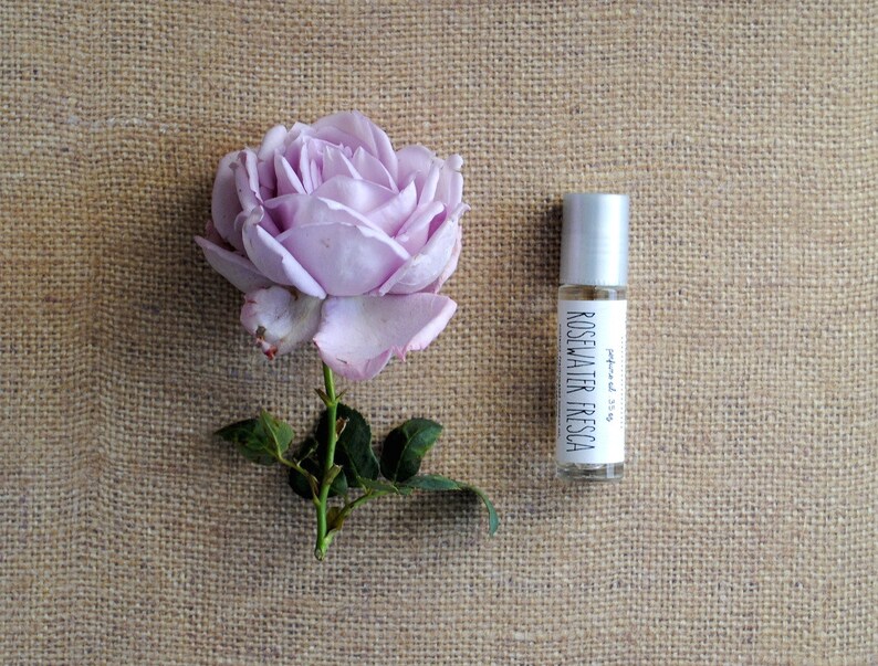 Op de afbeelding: Een klein glazen flesje met een zilveren rolbal bovenop. Het flesje is gelabeld "Rosewater Fresca" en "geparfumeerd 5 ml". Een enkele paarse roos met groene bladeren op een bruine jute achtergrond.