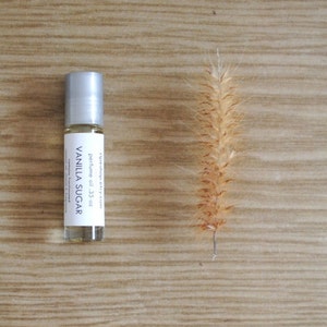 May include: A small glass bottle with a silver roller ball top. The bottle contains a clear liquid and has a white label that reads "raindrop city" and "perfume oil - .35 oz" in black text. The label also says "VANILLA SUGAR" in larger black text. The bottle is sitting on a light brown wooden surface.