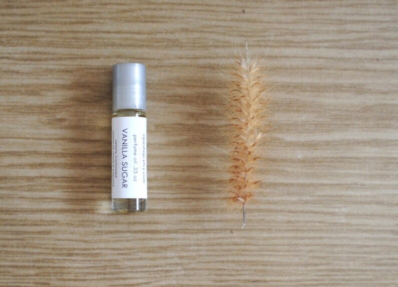 May include: A small glass bottle with a silver roller ball top. The bottle contains a clear liquid and has a white label that reads "raindrop city" and "perfume oil - .35 oz" in black text. The label also says "VANILLA SUGAR" in larger black text. The bottle is sitting on a light brown wooden surface.