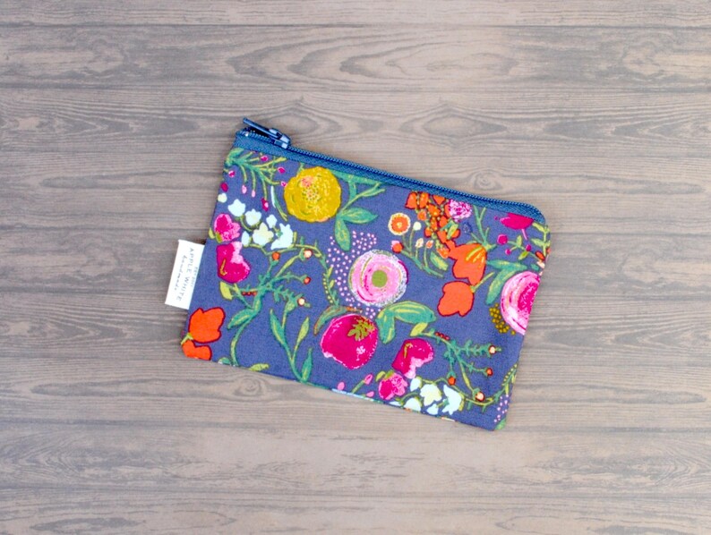 May include: A floral print pouch with a blue zipper. The pouch features a vibrant pattern of pink, orange, and yellow flowers on a blue background. A small white tag is attached to the side.