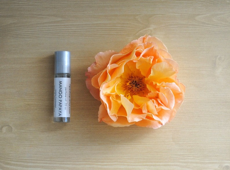 May include: A silver roller bottle with a white label that reads "mango papaya" and "perfume oil .33 oz". The bottle is sitting on a light brown wooden surface next to a peach-colored flower.