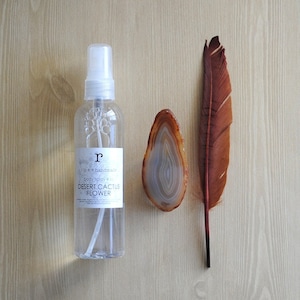 Pu&ograve; includere: Un flacone di vetro trasparente con un vaporizzatore bianco e un'etichetta che recita "ripe & handmade body spray + oil DESERT CACTUS FLOWER". Il flacone &egrave; appoggiato su una superficie di legno accanto a una piuma marrone e una fetta di agata.