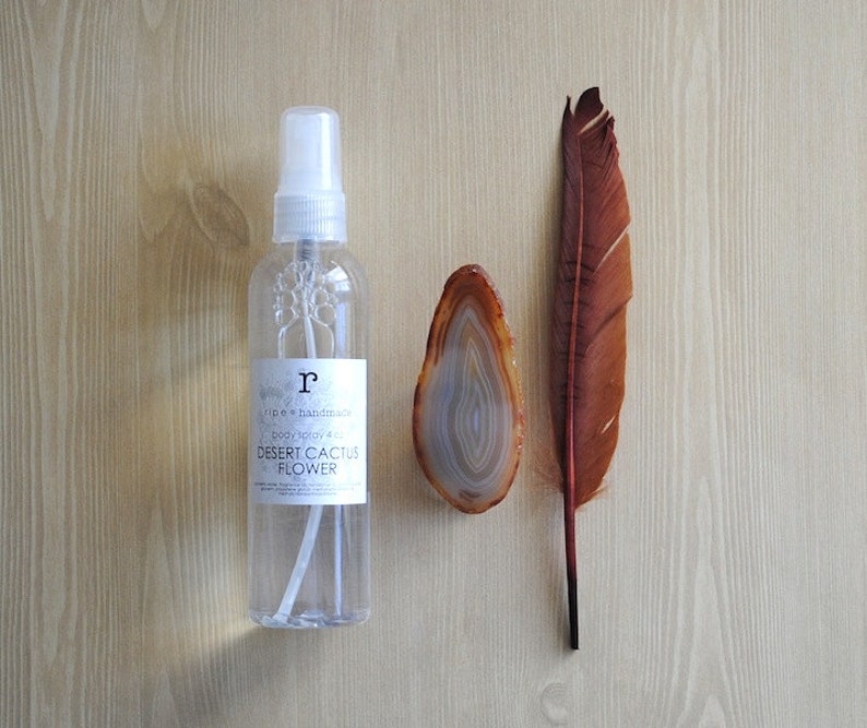 Pu&ograve; includere: Un flacone di vetro trasparente con un vaporizzatore bianco e un'etichetta che recita "ripe & handmade body spray + oil DESERT CACTUS FLOWER". Il flacone &egrave; appoggiato su una superficie di legno accanto a una piuma marrone e una fetta di agata.