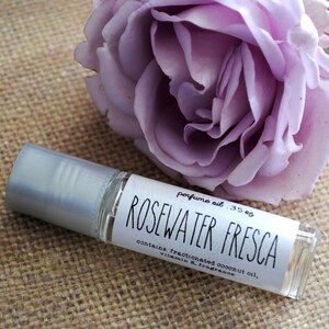 Op de afbeelding: Een klein glazen rolletje met een zilveren dop en een wit etiket met de tekst "perfume oil .35 oz ROSEWATER FRESCA contains: fractionated coconut oil, vitamin E, fragrance". De fles staat op een bruine jute ondergrond naast een paarse roos.