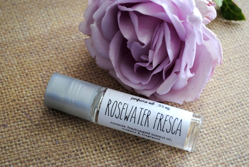 Op de afbeelding: Een klein glazen rolletje met een zilveren dop en een wit etiket met de tekst "perfume oil .35 oz ROSEWATER FRESCA contains: fractionated coconut oil, vitamin E, fragrance". De fles staat op een bruine jute ondergrond naast een paarse roos.