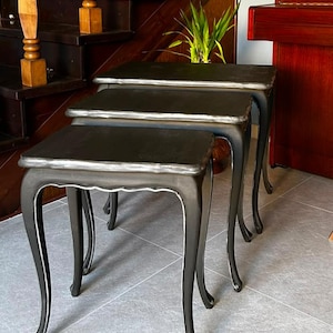 Può includere: Un set di tre tavolini a incastro con finitura nero opaco e bordi color argento anticato. Ogni tavolino presenta un piano rettangolare e quattro gambe curve con un dettaglio a grembiule smerlato, progettati per essere impilati.