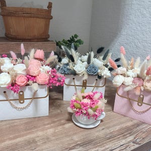 Può includere: Arrangiamenti floreali decorativi in scatole e una tazza da tè. I fiori sono bianchi, rosa e blu. Le scatole sono bianche e rosa con dettagli dorati e manici a catena. Un cestino di legno è sullo sfondo.