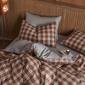May include: A bed dressed with a brown and white plaid duvet cover, pillows, and sheets. The pillows feature a brown, blue, and white plaid design. The sheets have a smaller, black and white checkered pattern. A small candle sits on a white tray.