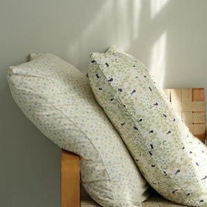 May include: Two long, patterned pillows rest on a wooden chair. One pillow features a cream background with small blue floral patterns. The other pillow has a cream background with green leaves and small black bird designs.
