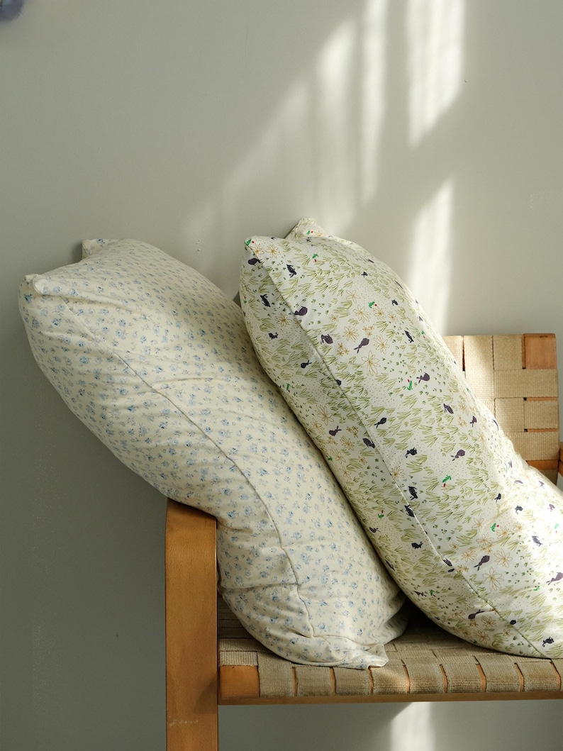 May include: Two long, patterned pillows rest on a wooden chair. One pillow features a cream background with small blue floral patterns. The other pillow has a cream background with green leaves and small black bird designs.