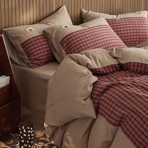 May include: Bed linen set in beige and red. The pillowcases have a beige base with a red plaid accent and a decorative embellishment. The duvet cover features a matching red plaid pattern. The bedding is arranged on a bed.