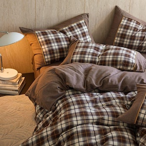 Puede incluir: Una cama con un edredón a cuadros marrón y blanco y fundas de almohada a juego. Las almohadas tienen un borde marrón. La cama está hecha con una sábana marrón y una funda nórdica a juego. La cama está en un dormitorio.