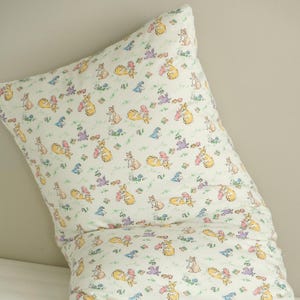 May include: A white pillow with a repeating pattern of bunnies in various poses, holding carrots and flowers. The bunnies are in shades of yellow, pink, and purple, with green grass and blue flowers.