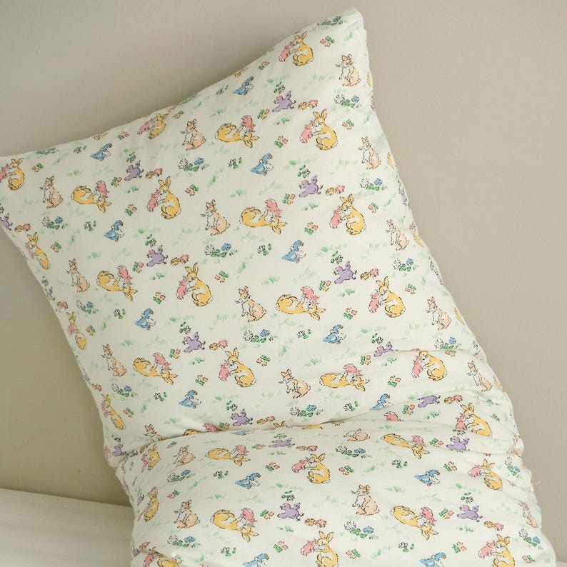 May include: A white pillow with a repeating pattern of bunnies in various poses, holding carrots and flowers. The bunnies are in shades of yellow, pink, and purple, with green grass and blue flowers.