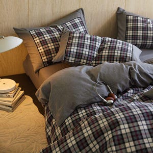 Può includere: Un letto con lenzuola e cuscini grigi. I cuscini e il copripiumino presentano un motivo a quadri nei toni del grigio, bianco e bordeaux. Un paio di cuffie poggia sul piumino. Una lampada e dei libri sono visibili a sinistra.