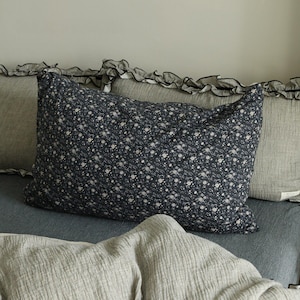 May include: A navy blue pillow with a white floral pattern is the focal point of this image. The pillow is surrounded by other pillows and bedding in shades of gray and blue. The bedding has a ruffled edge.