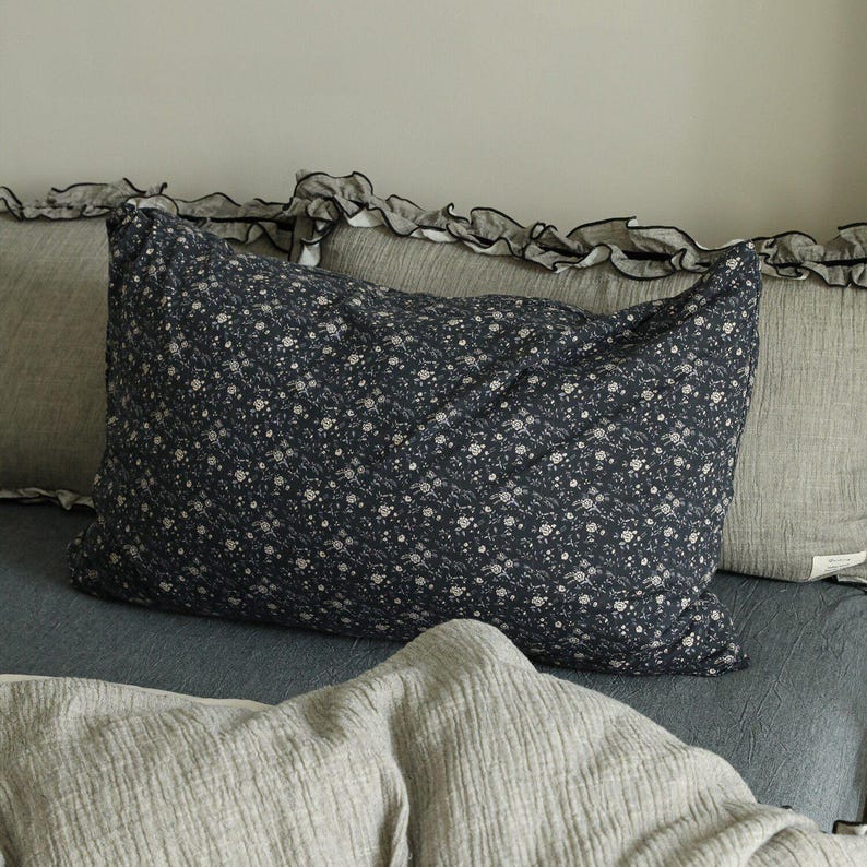 May include: A navy blue pillow with a white floral pattern is the focal point of this image. The pillow is surrounded by other pillows and bedding in shades of gray and blue. The bedding has a ruffled edge.
