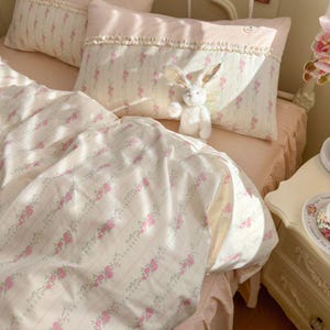 Könnte beinhalten: Ein Bett mit floral gemusterter Bettwäsche in Creme und Rosa. Die Bettwäsche umfasst einen Bettbezug, Kissenbezüge und ein dekoratives Kissen mit Rüschenbesatz. Ein weißes Stoffkaninchen sitzt auf den Kissen. Das Gesamtthema ist ein Vintage-Schlafzimmer im romantischen Stil.
