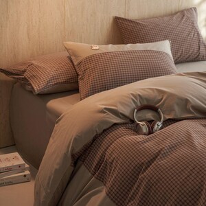 Pode incluir: Uma cama com uma colcha e fronhas xadrez marrons e brancas. A cama também tem um lençol marrom liso e um par de fones de ouvido repousando sobre a colcha. A roupa de cama está bem arrumada, sugerindo um ambiente de quarto confortável e convidativo.
