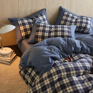 May include: A blue plaid bedding set with matching pillowcases and a duvet cover. The pillows have a dark blue border. A pair of headphones rests on the bed. A small lamp and books are on a nightstand.