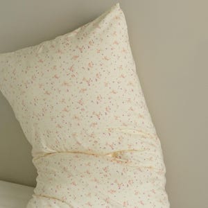 May include: A cream-colored pillow with a delicate floral pattern in shades of pink and purple. The pillow is rectangular and appears to be made of soft fabric, perfect for a bedroom or living room setting.