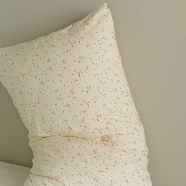 May include: A cream-colored pillow with a delicate floral pattern in shades of pink and purple. The pillow is rectangular and appears to be made of soft fabric, perfect for a bedroom or living room setting.