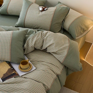 Può includere: Set di biancheria da letto verde salvia con cuscini fantasia e una striscia rossa. Una tazza di caffè e un libro sono sul letto. Le cuffie sono appoggiate sui cuscini.